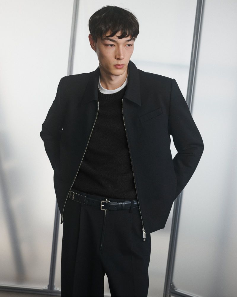 Portrait of a young adult man in a black jacket, sweater, and pants against a frosted glass background in a studio setting.