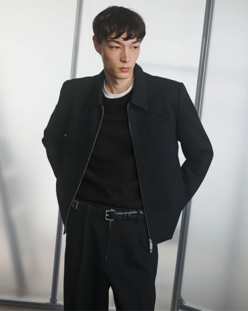 Portrait of a young adult man in a black jacket, sweater, and pants against a frosted glass background in a studio setting.