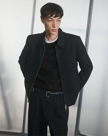 Portrait of a young adult man in a black jacket, sweater, and pants against a frosted glass background in a studio setting.