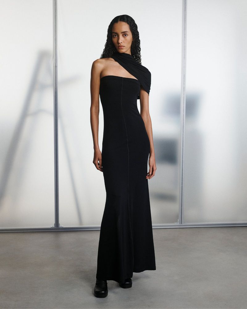 A slender woman in a black, one-shoulder gown poses elegantly against a neutral background, highlighting the gown's modern design.