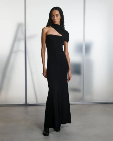 A slender woman in a black, one-shoulder gown poses elegantly against a neutral background, highlighting the gown's modern design.