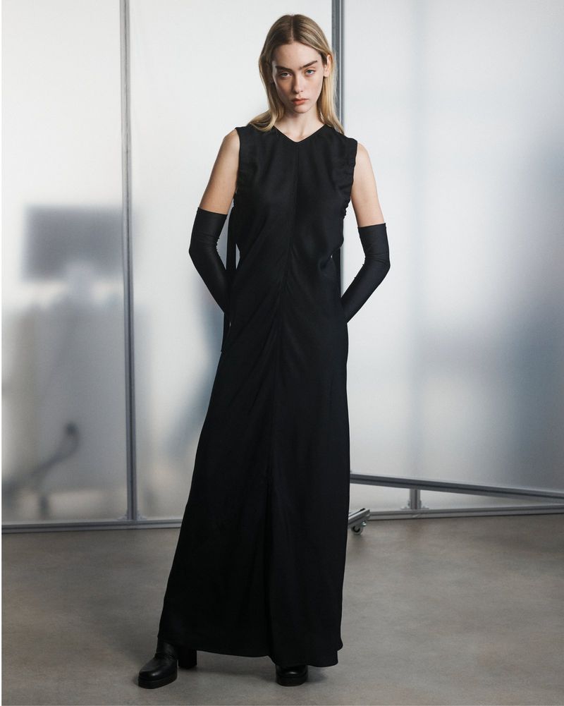 Fashion model in a sleeveless black maxi dress with long black gloves posing in front of a frosted glass panel backdrop in a studio.