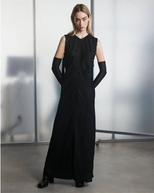 Fashion model in a sleeveless black maxi dress with long black gloves posing in front of a frosted glass panel backdrop in a studio.