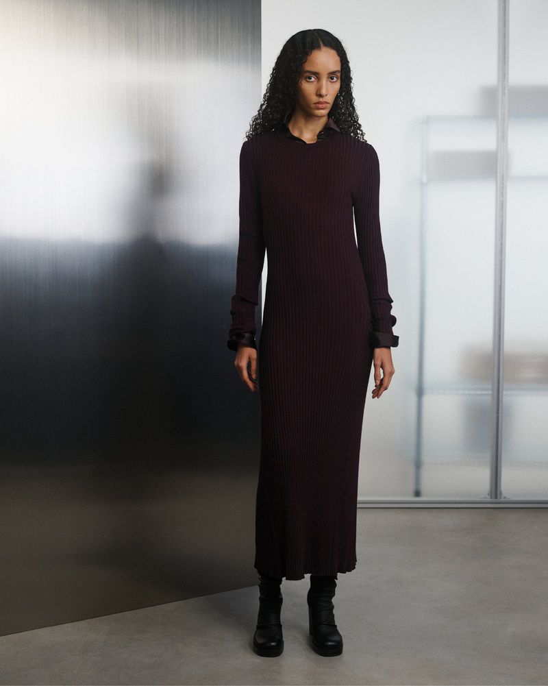 A woman with curly hair poses in a long-sleeve, ribbed burgundy dress and black boots against a modern, minimalist background indoors.