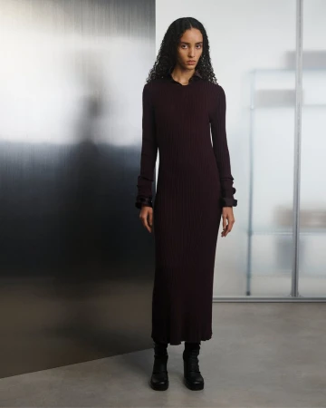 A woman with curly hair poses in a long-sleeve, ribbed burgundy dress and black boots against a modern, minimalist background indoors.