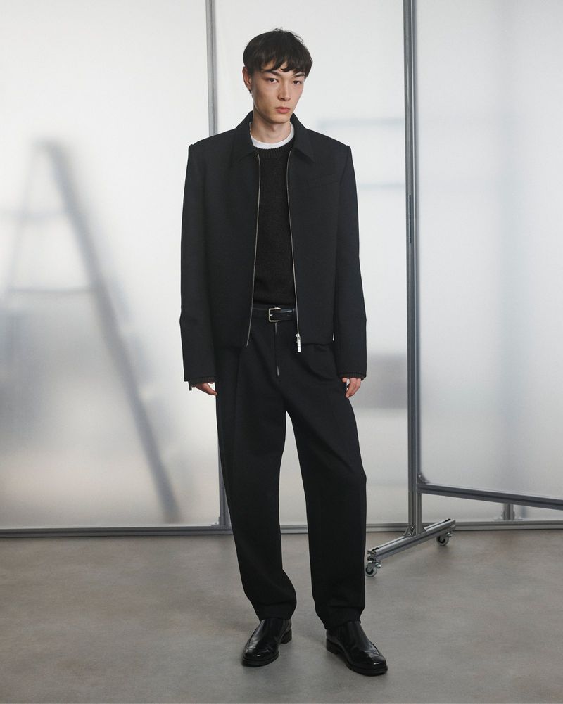 A full-length portrait captures a young adult male fashion model wearing a black jacket, black shirt, and black trousers indoors.