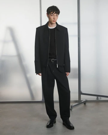 A full-length portrait captures a young adult male fashion model wearing a black jacket, black shirt, and black trousers indoors.