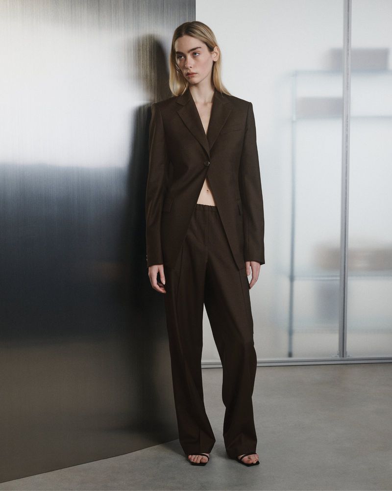 A model poses in a brown suit in a minimalist studio. The composition features clean lines, neutral colors, and natural light, showcasing contemporary fashion.