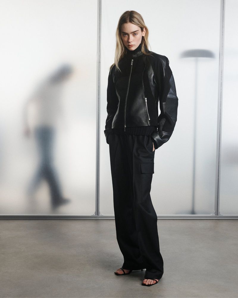 Fashion model in black leather jacket and wide-leg trousers poses in a studio with frosted glass backdrop. Full-length shot, neutral expression.