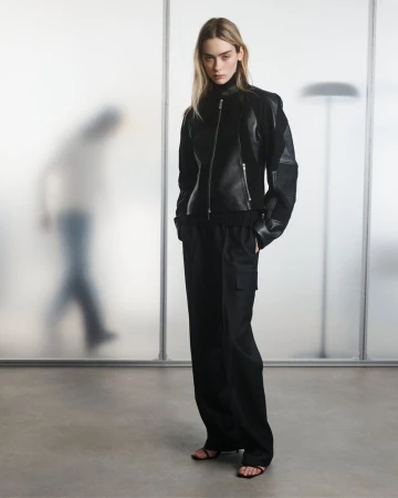 Fashion model in black leather jacket and wide-leg trousers poses in a studio with frosted glass backdrop. Full-length shot, neutral expression.