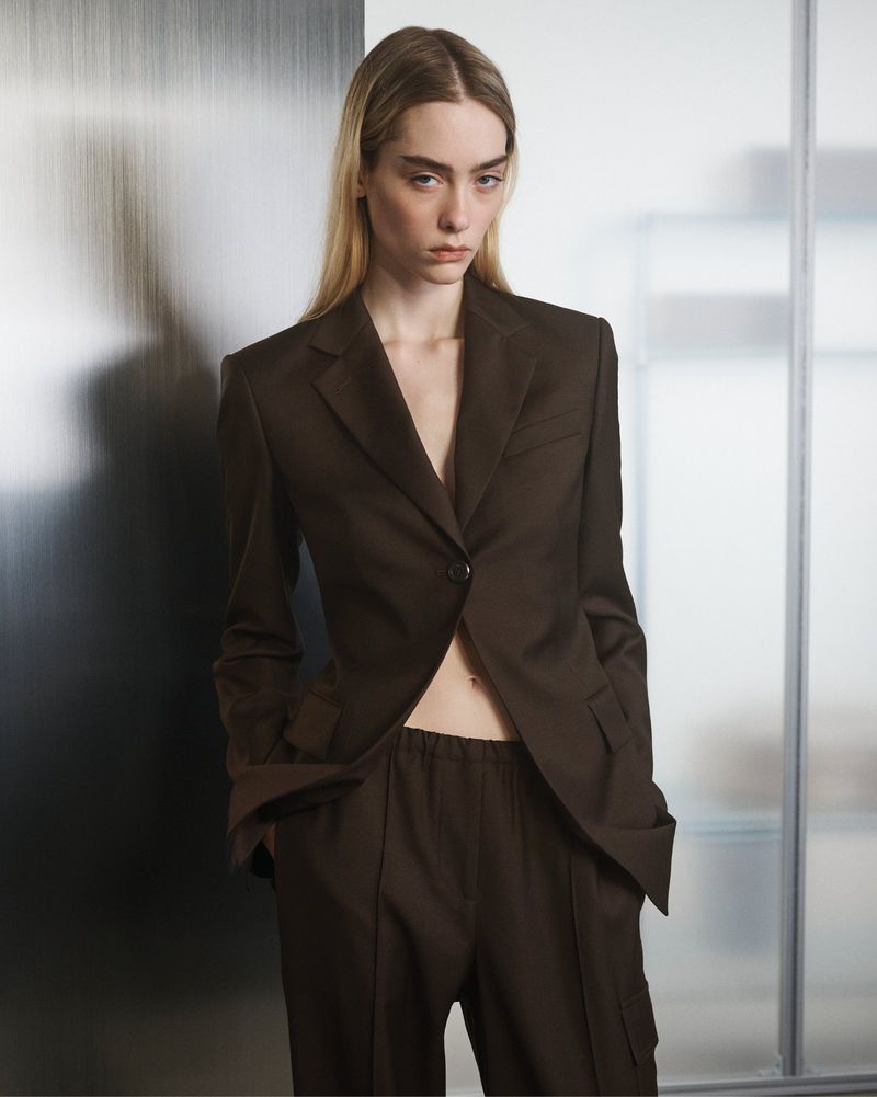 Blonde woman in a brown blazer and matching pants poses in front of a textured wall, showcasing a minimalist fashion aesthetic.