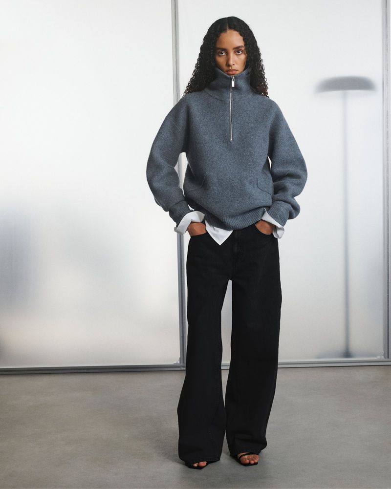 Studio shot of a woman in a grey sweater and black pants, hands in pockets, against a blurred background with translucent panels.