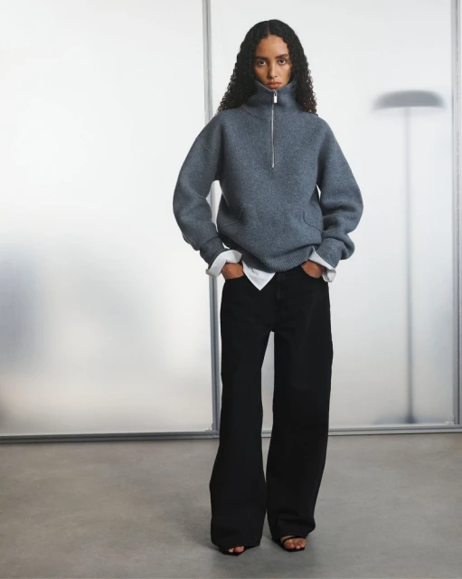 Studio shot of a woman in a grey sweater and black pants, hands in pockets, against a blurred background with translucent panels.