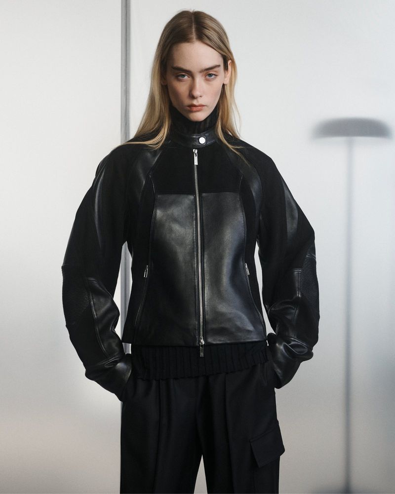 Model wearing a black mixed-media leather and knit zip jacket with black tailored pants in front of a diffused grey studio backdrop.