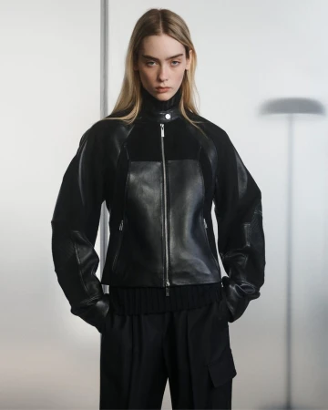 Model wearing a black mixed-media leather and knit zip jacket with black tailored pants in front of a diffused grey studio backdrop.