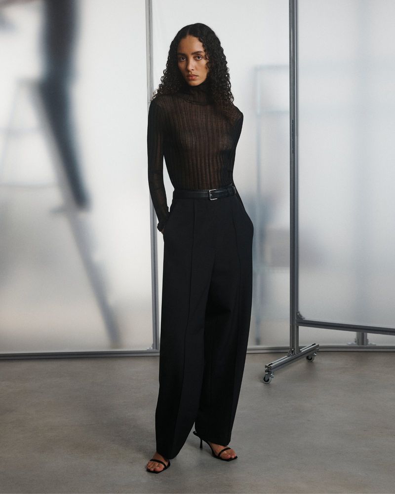 A woman with curly hair poses in a black turtleneck and wide-leg pants, standing against a frosted glass pane in a studio setting.