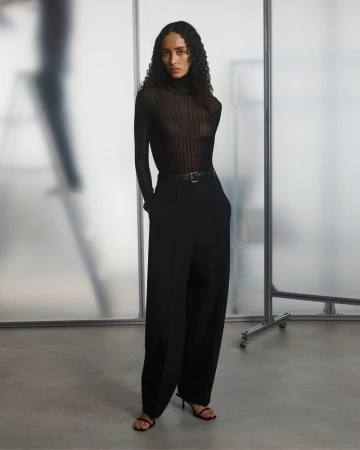 A woman with curly hair poses in a black turtleneck and wide-leg pants, standing against a frosted glass pane in a studio setting.
