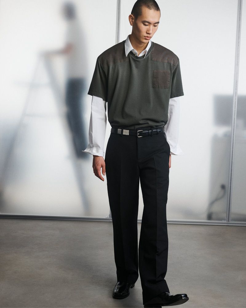A full-length portrait of a young adult male in contemporary fashion, standing in front of a frosted-glass paneled wall in a photo studio.