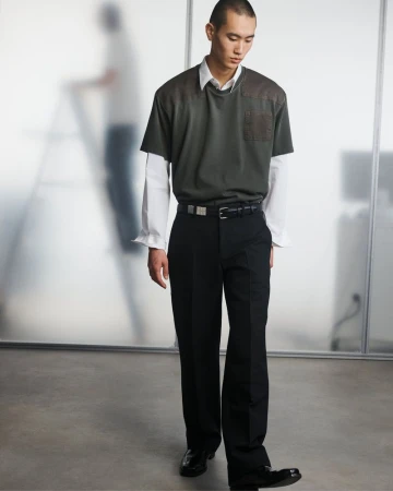 A full-length portrait of a young adult male in contemporary fashion, standing in front of a frosted-glass paneled wall in a photo studio.
