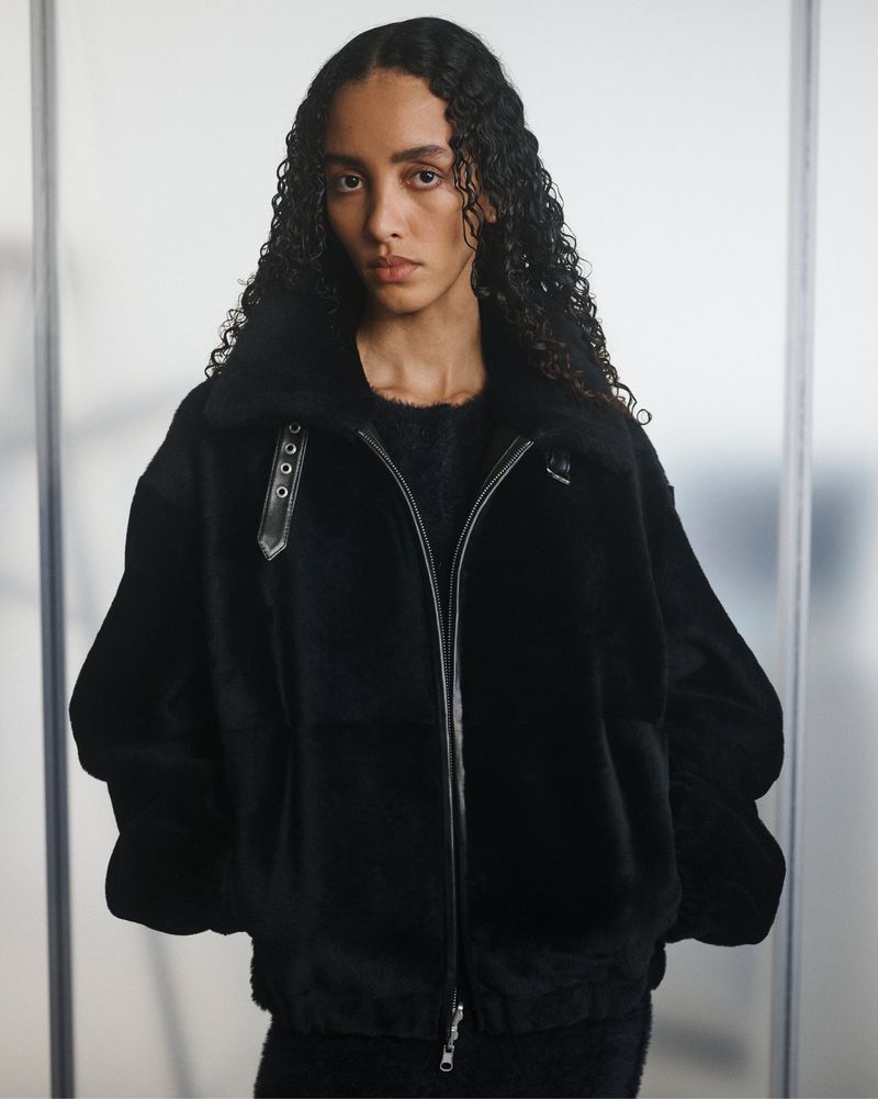 A woman with curly hair poses in a black faux fur jacket with metal hardware against a neutral background, creating a fashionable and cozy look.