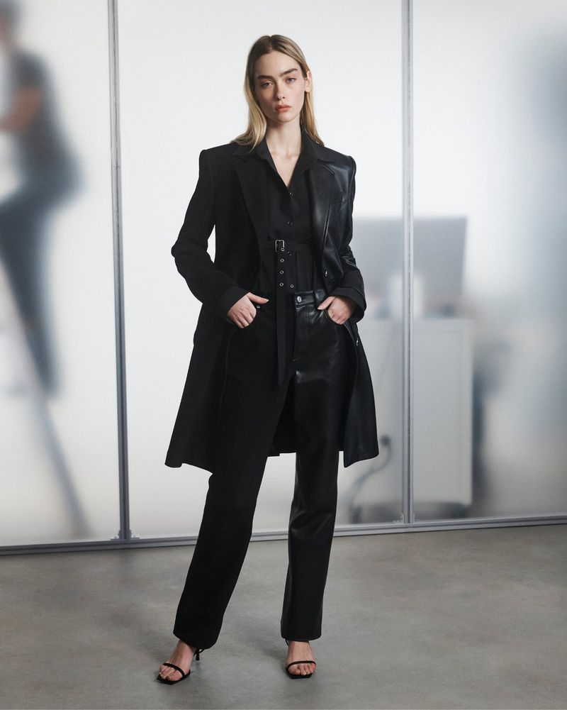 A fashionable woman in a black coat and pants stands in front of a glass wall in a minimalist style, illuminated by soft, diffused light.