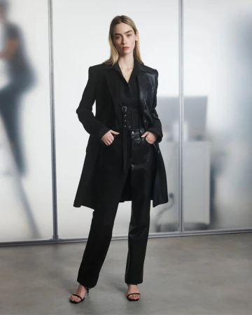 A fashionable woman in a black coat and pants stands in front of a glass wall in a minimalist style, illuminated by soft, diffused light.