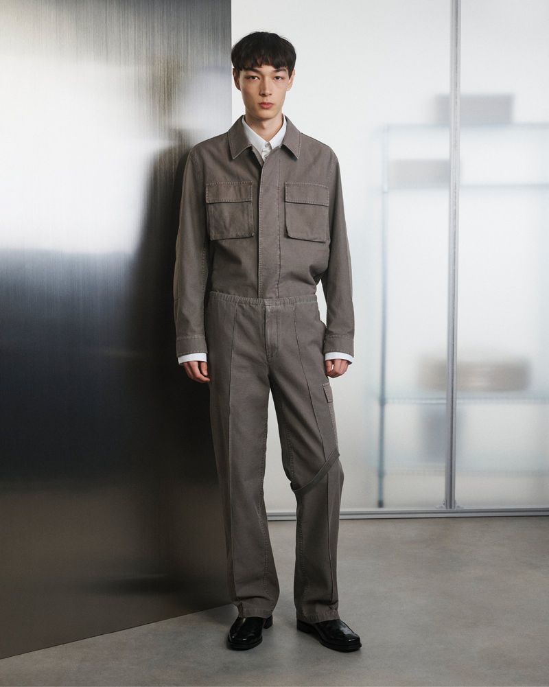 Full-length studio shot of a young adult male model wearing a grey jumpsuit and black leather shoes, posing against a grey and white backdrop.