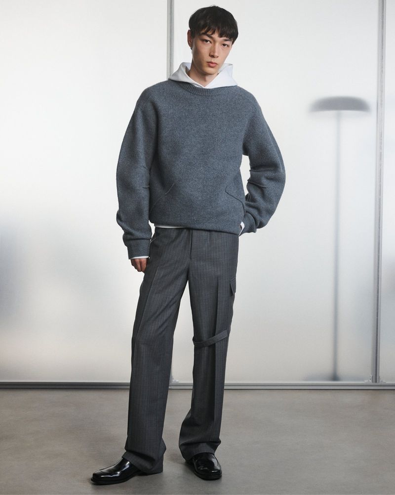 Studio shot of a young male model wearing a grey sweatshirt, pinstripe dress pants and black dress shoes, posing against a grey background.