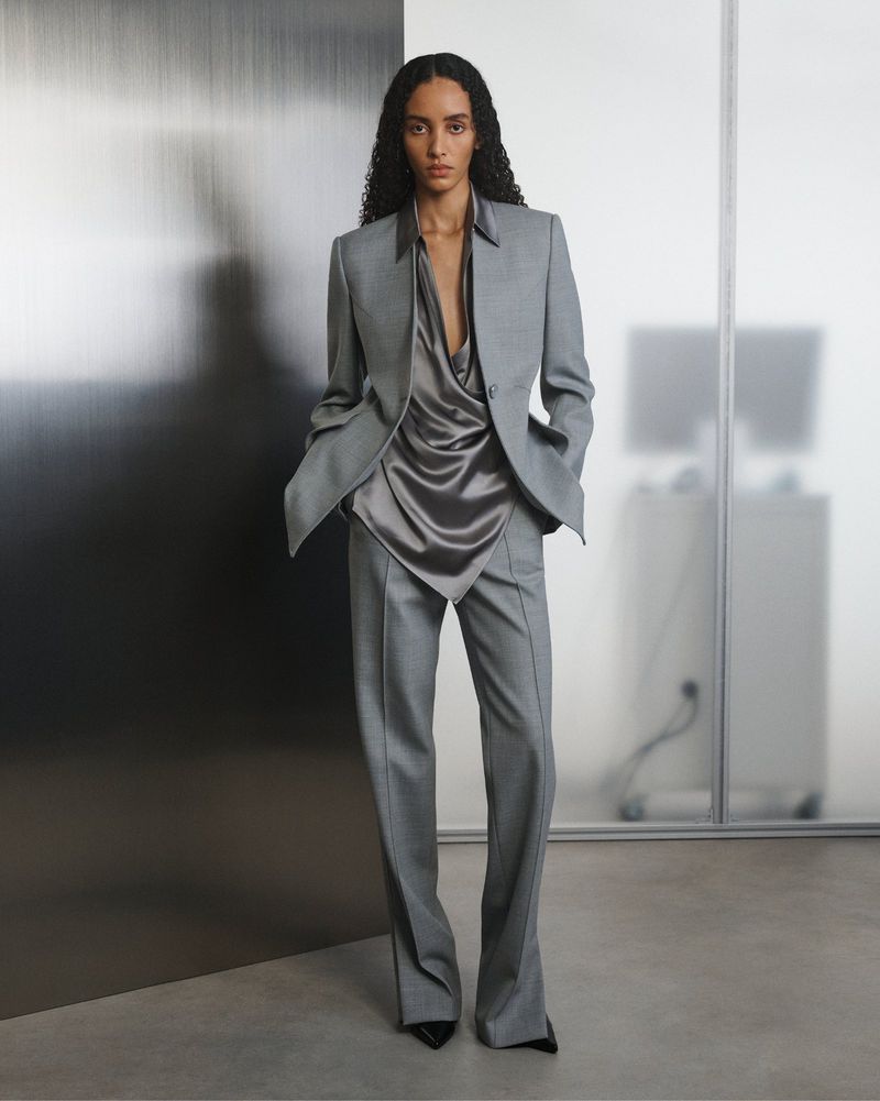 A woman wearing a grey suit and a silky top standing in a studio. Fashion shot with a full shot and eye-level camera angle.