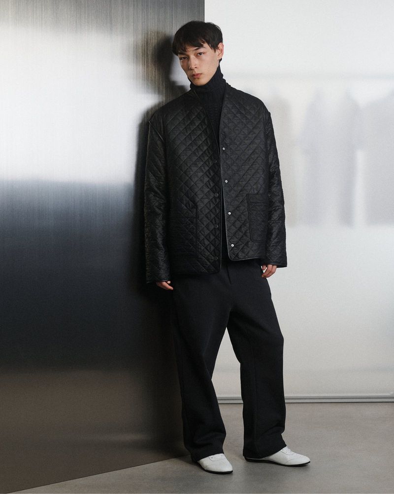 A slender Asian man stands indoors wearing a black turtleneck, quilted jacket, black pants, and white shoes, posing against a gradient wall.