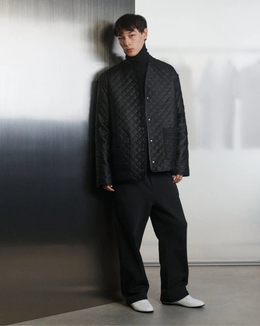 A slender Asian man stands indoors wearing a black turtleneck, quilted jacket, black pants, and white shoes, posing against a gradient wall.