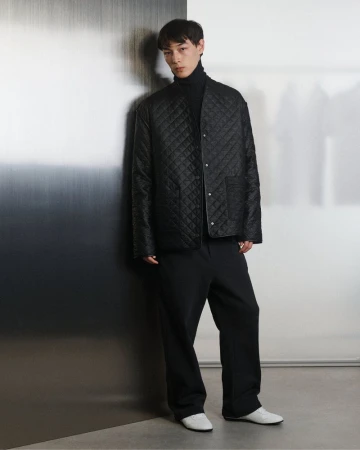 A slender Asian man stands indoors wearing a black turtleneck, quilted jacket, black pants, and white shoes, posing against a gradient wall.