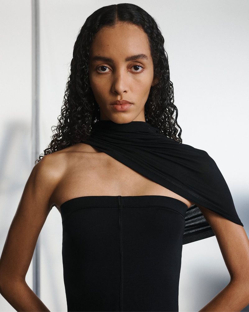 Portrait of a woman with dark curly hair wearing a black, one-shoulder garment, looking directly at the camera against a white backdrop.