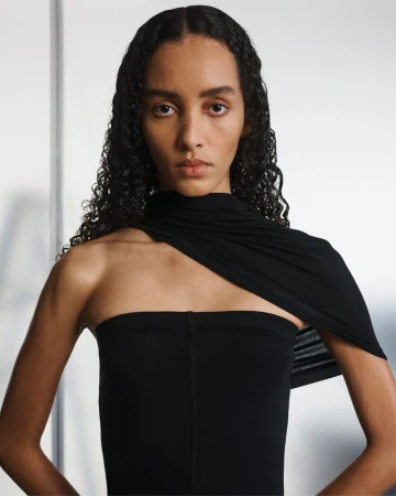 Portrait of a woman with dark curly hair wearing a black, one-shoulder garment, looking directly at the camera against a white backdrop.