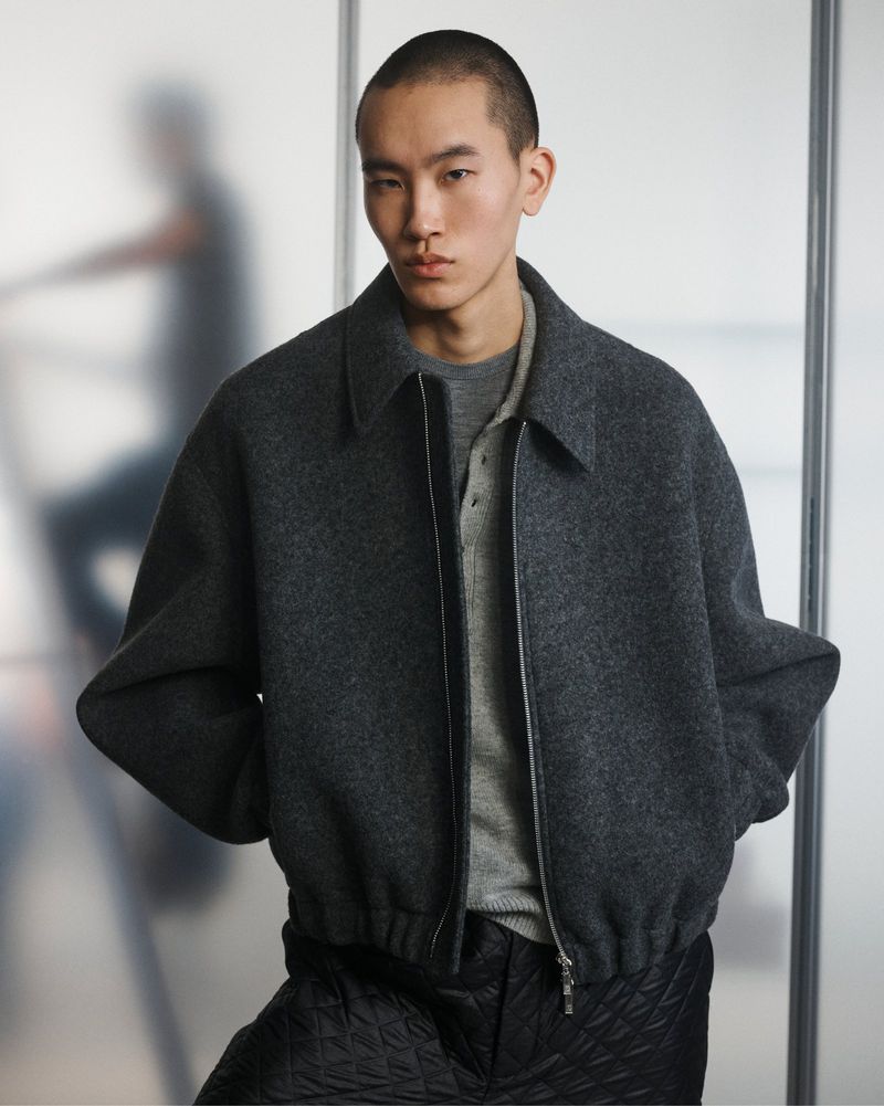 Adult male poses in a grey wool bomber jacket and quilted black pants in an indoor photo studio setting, showcasing modern fashion.