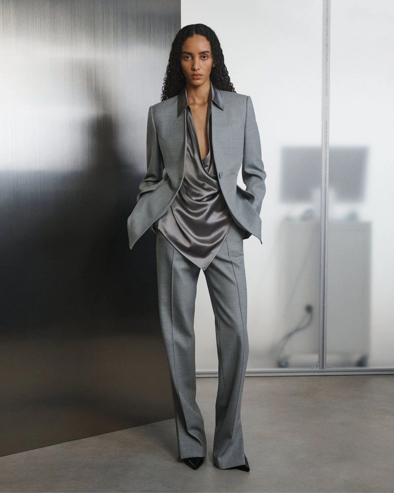Model posing in a tailored grey suit and draped satin blouse against a brushed metal and frosted glass partition.
