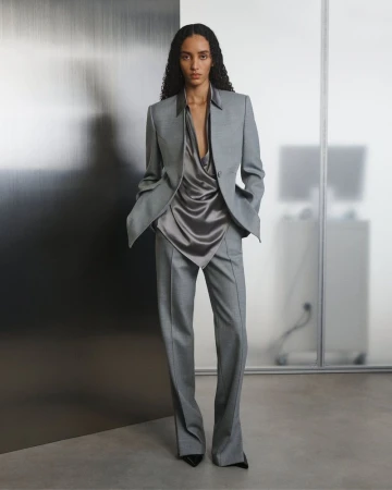 Model posing in a tailored grey suit and draped satin blouse against a brushed metal and frosted glass partition.