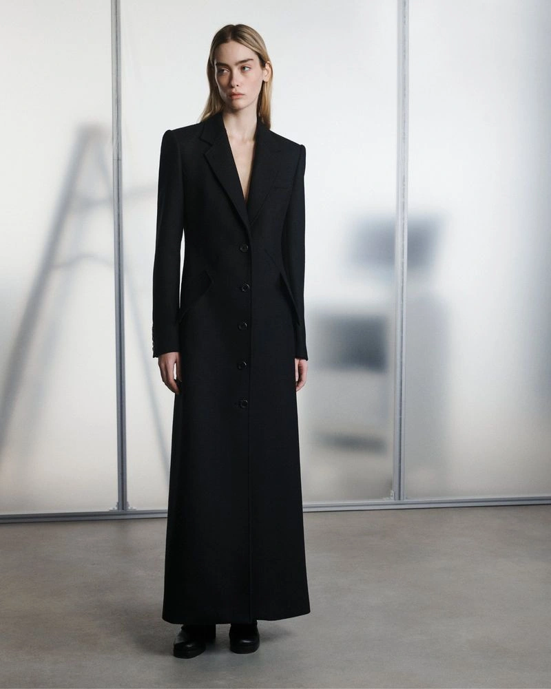Model wearing a floor-length black tailored coat standing in front of a translucent studio backdrop.