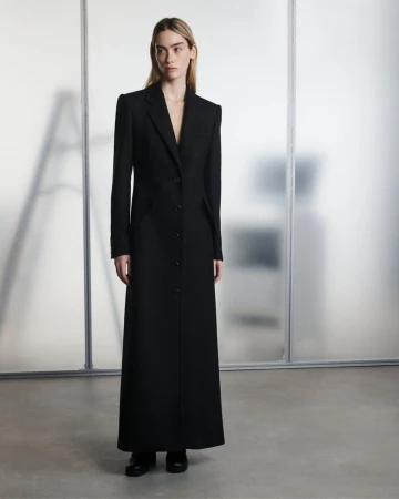 Model wearing a floor-length black tailored coat standing in front of a translucent studio backdrop.