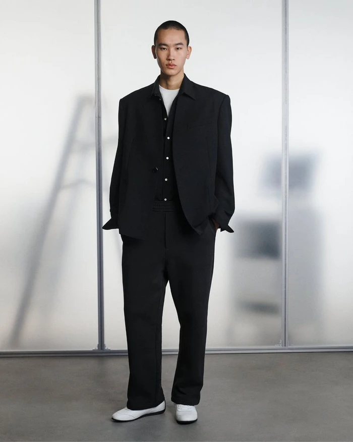 A man wearing a black suit jacket and trousers over a white t-shirt standing against a pale translucent background.