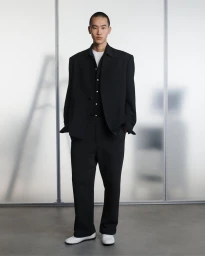 A man wearing a black suit jacket and trousers over a white t-shirt standing against a pale translucent background.