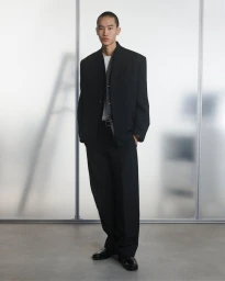 Model wearing an oversized black suit jacket and wide-leg trousers standing before frosted glass studio panels.