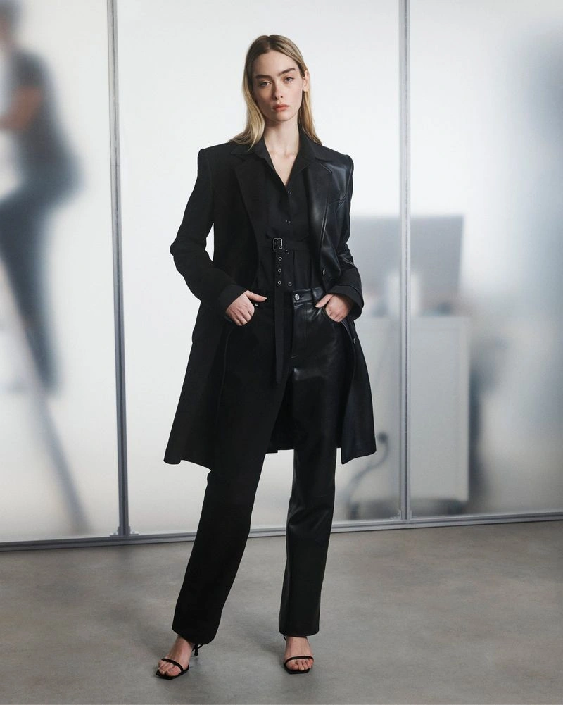 Model wearing a tailored black coat, black leather trousers, and strappy heels against frosted glass studio panels.