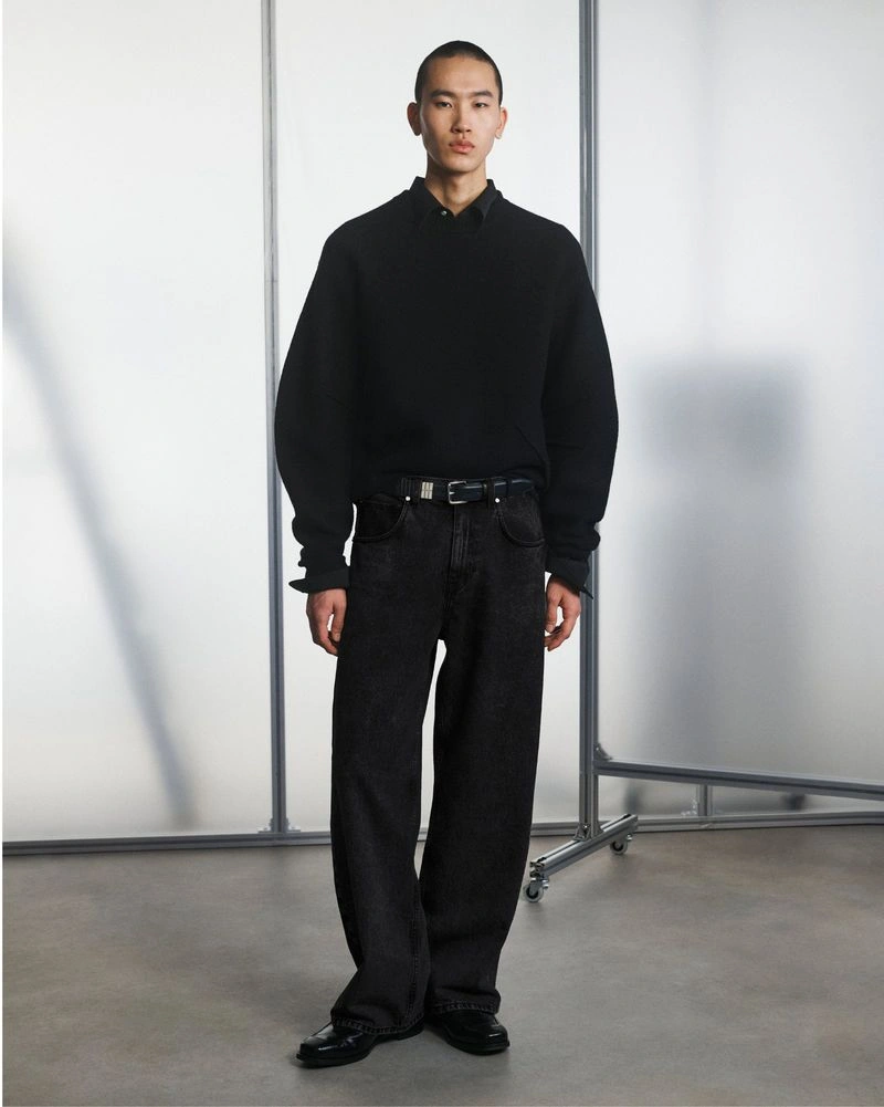 East Asian male model posing in a black crewneck sweater, collared shirt, and relaxed dark jeans against frosted studio screens.