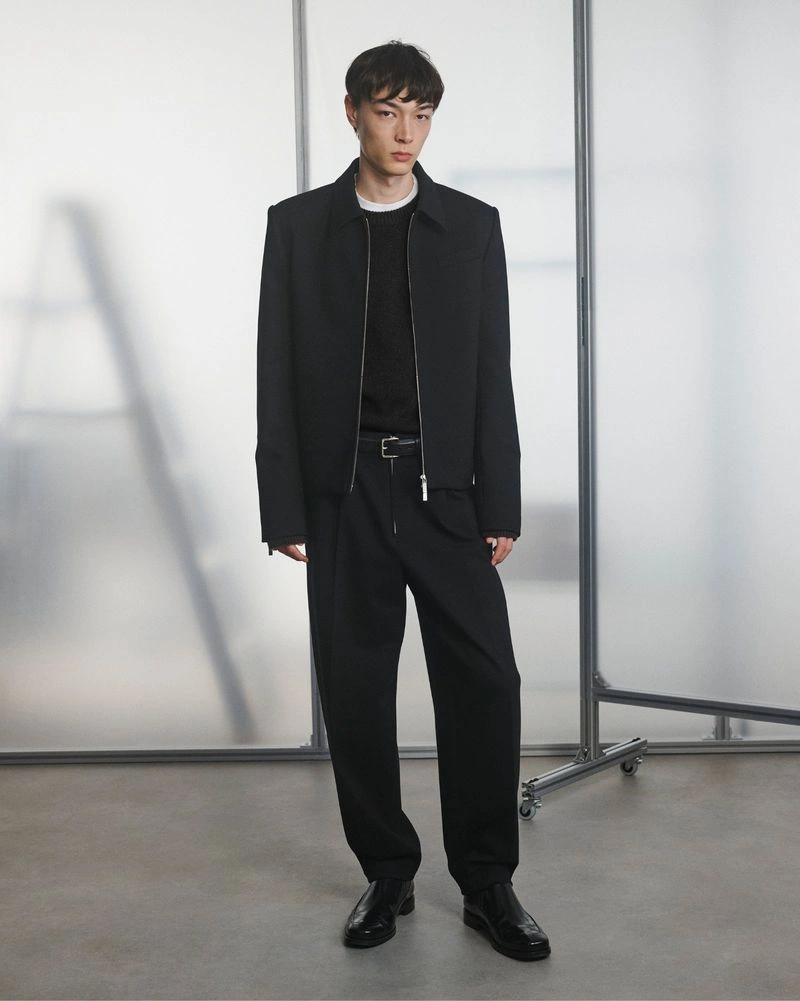 Model wearing a black zip-up jacket and matching relaxed trousers in front of frosted studio screens for Helmut Lang.