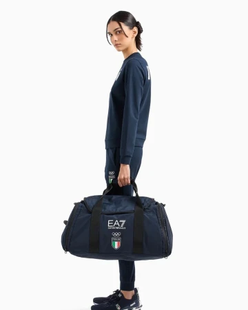 A full-length shot showcases a female model holding a navy blue duffel bag, dressed in a matching tracksuit against a white background.