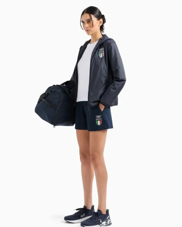 A woman in sport clothes stands holding a duffel bag. She wears a jacket, shorts, and sneakers, all with the Italian flag logo.