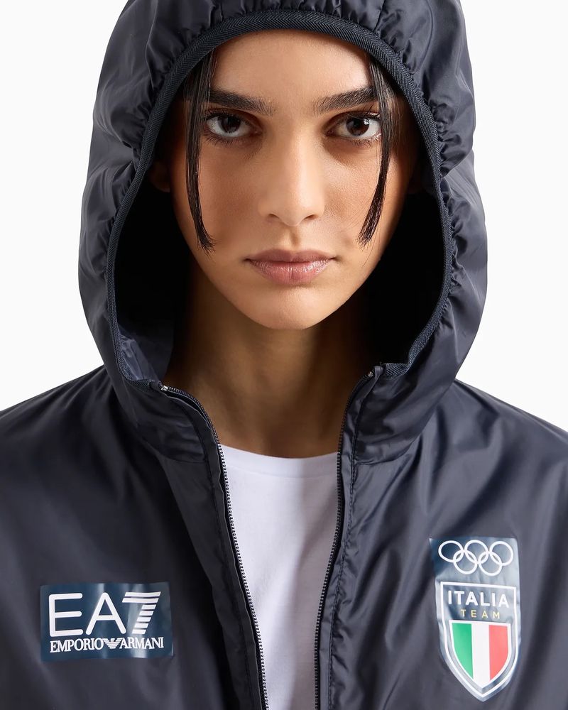 Close-up of a woman wearing a hooded blue Emporio Armani windbreaker with Italia Team logo, looking directly at the camera on white.