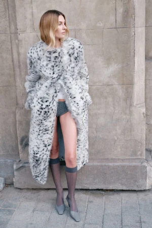 Woman in a long, spotted faux-fur coat poses against a textured wall, highlighting fashion, style, and urban ambiance with sheer hosiery.