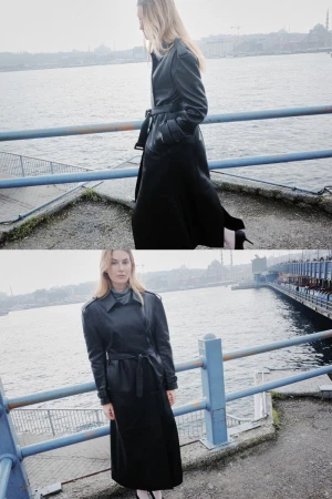 An adult woman in a long black coat poses near a waterway, city visible in the distance during daytime, with soft, diffused lighting.